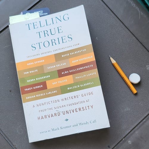 The book Telling True Stories: A Nonfiction Writer's Guide from the Neiman Foundation at Harvard University sits on an outdoor table. Post-it notes and a book mark stick out of the top; a short pencil and a white eraser sit next to it.